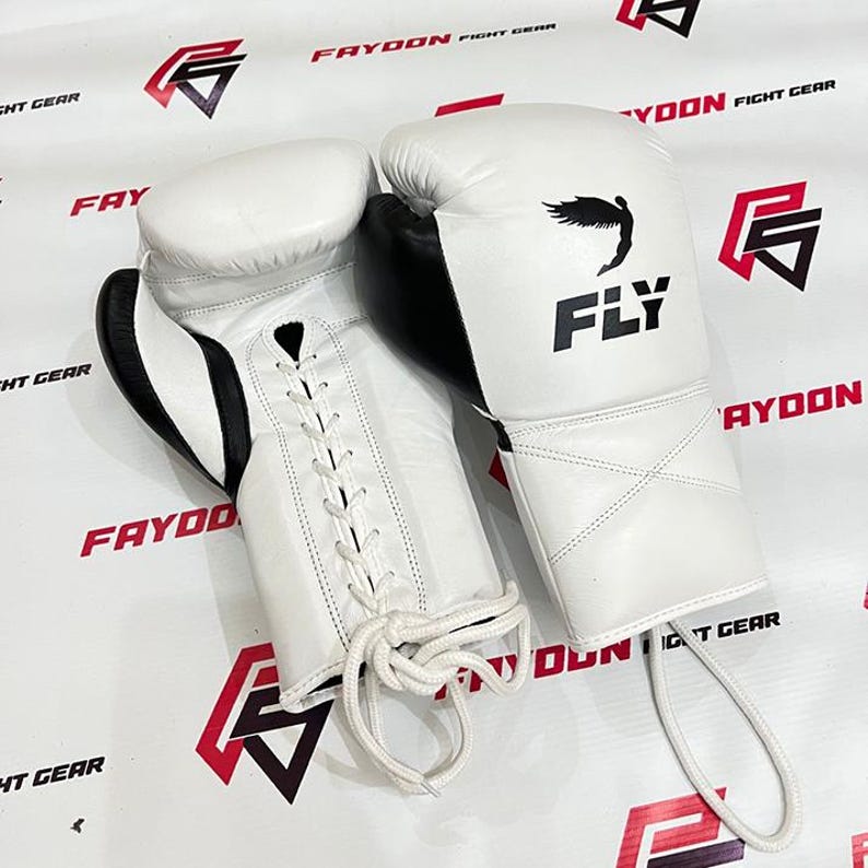May include: A pair of white boxing gloves with black detailing. The gloves feature a lace-up closure and the word "FLY" is printed on one glove, along with a bird graphic. These gloves are designed for combat sports.