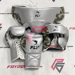 May include: A silver boxing set featuring a head guard, gloves, and groin protector. The set has camouflage accents and the "FLY" logo. The gloves are laced up with white laces. The set is designed for boxing and other combat sports.