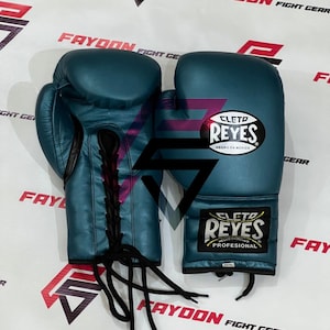 以下が含まれることがあります： 黒い紐が付いた、ティール色のボクシンググローブのペア。「CLETO REYES」のロゴが白と金で表示されています。メキシコ製。プロ仕様です。