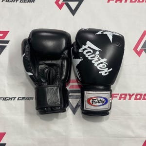 Puede incluir: Un par de guantes de boxeo negros con el logotipo blanco de Fairtex impreso en la parte frontal y un parche rectangular con el nombre de la marca Fairtex en rojo, blanco y azul en la muñequera. Los guantes tienen un cierre de velcro.