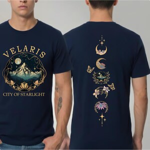 May include: Navy blue t-shirt with the word "VELARIS" above a mountain scene and the text "CITY OF STARLIGHT". The back of the shirt features a design of moon phases, butterflies, and floral elements in gold.