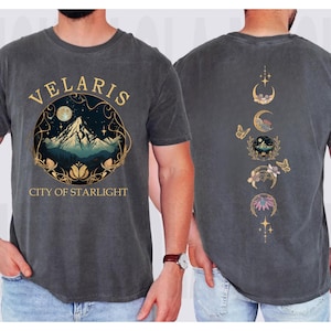 May include: Dark gray t-shirt with a front graphic featuring the text "VELARIS CITY OF STARLIGHT" around a mountain scene with a moon and stars. The back of the shirt has a design of moon phases, butterflies, and floral elements.