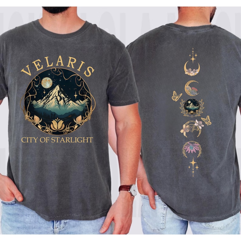 May include: Dark gray t-shirt with a front graphic featuring the text "VELARIS CITY OF STARLIGHT" around a mountain scene with a moon and stars. The back of the shirt has a design of moon phases, butterflies, and floral elements.