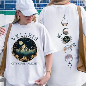 May include: White t-shirt with the text "VELARIS CITY OF STARLIGHT" and a mountain and moon graphic. The back of the shirt features a design of moon phases, butterflies, and floral elements. The shirt is a casual, comfortable style.
