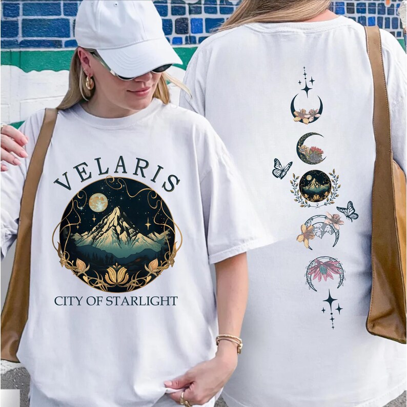 May include: White t-shirt with the text "VELARIS CITY OF STARLIGHT" and a mountain and moon graphic. The back of the shirt features a design of moon phases, butterflies, and floral elements. The shirt is a casual, comfortable style.