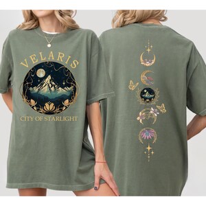 May include: Sage green t-shirt with a front graphic featuring the text "VELARIS CITY OF STARLIGHT" around a mountain scene. The back of the shirt has a design of moon phases, butterflies, and floral elements. The shirt is made of a soft material.