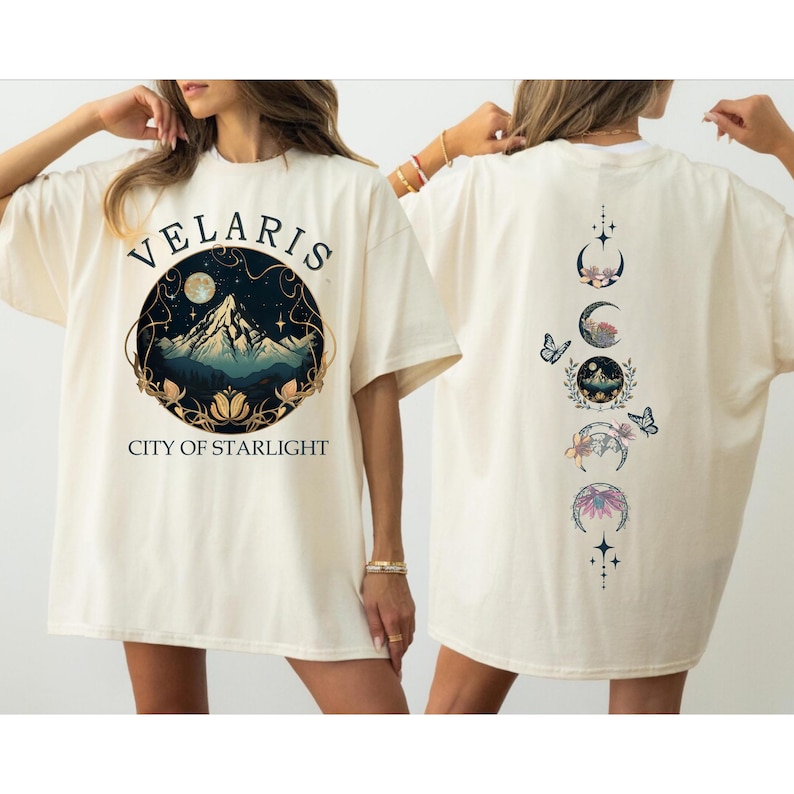 May include: Cream-colored t-shirt featuring the text "VELARIS CITY OF STARLIGHT" above a mountain and moon graphic. The back of the shirt displays a vertical design of moon phases, butterflies, and floral elements. The shirt has short sleeves and a relaxed fit.