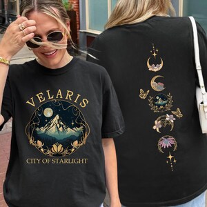 May include: Black t-shirt with the text "VELARIS" above a mountain and moon graphic. The back of the shirt features a design of moons, butterflies, and floral elements. The shirt is a casual wear item.