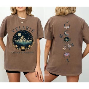 May include: Brown t-shirt with the text "VELARIS CITY OF STARLIGHT" in a circular design featuring a mountain and moon. The back of the shirt has a vertical design of moon phases, flowers, and butterflies.