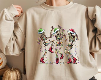 May include: Beige sweatshirt featuring a festive design of three dancing skeletons wearing Santa hats and red boots. The skeletons are adorned with colorful Christmas lights, creating a whimsical holiday theme. The sweatshirt is a neutral color.