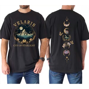 May include: Black t-shirt with "VELARIS CITY OF STARLIGHT" text and a mountain scene graphic on the front. The back features a design of crescent moons, butterflies, and floral elements in gold and other colors.