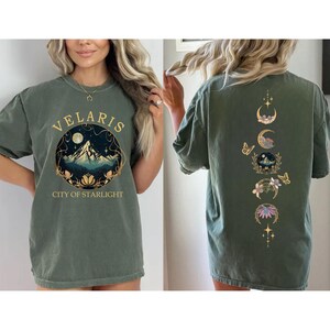 May include: Olive green t-shirt with the word "VELARIS" above a mountain and starry night design. The back of the shirt features a vertical design of moon phases, flowers, and butterflies in gold.