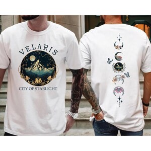 May include: White t-shirts with a graphic design. One shirt features the text "VELARIS CITY OF STARLIGHT" with a mountain and moon design. The other shirt has a design of moon phases and butterflies.