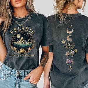 May include: Dark gray t-shirt with "VELARIS CITY OF STARLIGHT" text and a mountain scene graphic on the front. The back features a design of moon phases, butterflies, and floral elements. The shirt is made of a soft material.
