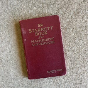 May include: A maroon hardcover book titled The Starrett Book for Machinists' Apprentices, Volume I. The front cover features gold embossed text with the title and publisher information, showing signs of wear on the edges and surface.