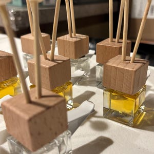 Könnte beinhalten: Eine Sammlung kleiner, quadratischer Glasflaschen, gefüllt mit gelber Flüssigkeit, gekrönt von Holzblöcken und dekorativen Holzstäben. Die Holzstäbe haben ein rundes, geschnitztes Design. Die Flaschen sind auf einer weißen Oberfläche angeordnet.