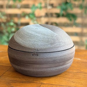 Könnte beinhalten: Ein runder Keramikbehälter mit Deckel und strukturierter Oberfläche. Der Behälter ist hauptsächlich braun mit einem grauen Abschnitt oben. Der Deckel hat eine dünne schwarze Linie am Rand. Das Wort "XUNTA" ist auf der Seite aufgedruckt.