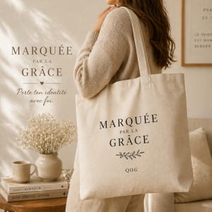 May include: A beige tote bag with the words "MARQUÉE PAR LA GRÂCE" printed in black. The bag also features a sprig design and the letters "QOG". The bag is being held by a person.