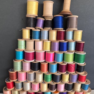 May include: A collection of vintage wooden spools of thread in various colors, stacked in a pyramid shape. The spools display a range of colors, including yellow, black, blue, pink, and green. The spools are arranged on a white surface against a dark gray background.