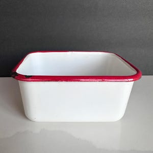 May include: A white enamel rectangular container with a vibrant red rim. The vintage-style container has a simple, clean design, perfect for kitchen or home decor. The container is empty and ready for use.