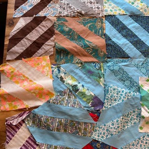 May include: A collection of colorful fabric squares with diagonal stripes. The squares feature various patterns, including floral prints, paisley, and solid colors in shades of blue, brown, pink, and purple. These fabric pieces are likely for quilting or other textile projects.