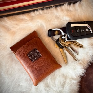 Op de afbeelding: Een bruine leren kaarthouder met een decoratief metalen embleem, naast een set sleutels en een autosleutel. De items liggen op een witte, zachte ondergrond, met een stapel boeken op de achtergrond.
