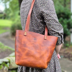 May include: A brown leather tote bag with a shoulder strap. The bag has a rectangular shape with a slightly textured surface. The bag is held by a person wearing a floral print dress. The bag has brass rivets and a small embossed logo.