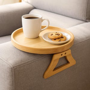 Puede incluir: Una bandeja de brazo de sofá de bambú con una taza blanca de café y un plato pequeño con dos galletas con chispas de chocolate. La bandeja tiene un diseño redondo y un accesorio de reposabrazos de madera.