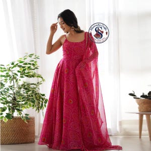 May include: A vibrant pink dress with a matching sheer scarf. The dress features a spaghetti strap neckline and a flowing, floor-length skirt. The fabric is patterned with a repeating design in shades of orange and yellow. The dress is suitable for special occasions.