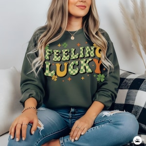 Pode incluir: Camisola verde escuro com o texto "FEELING LUCKY" em uma fonte retrô. As letras são uma mistura de verde xadrez, amarelo e laranja. O design inclui trevos e estrelas, perfeito para o Dia de São Patrício.