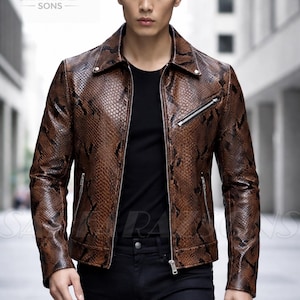 Może przedstawiać: Brązowa skórzana kurtka z wzorem skóry węża i srebrnym zapięciem na zamek błyskawiczny. Kurtka ma klasyczny kołnierz i dwie kieszenie zapinane na zamek. Model ma na sobie czarną koszulę i czarne spodnie. Widoczna jest nazwa marki "SARFARAZ SONS".