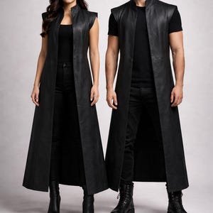 May include: A long, sleeveless black leather vest featuring a high collar and an open front design. The vest extends to the mid-calf and is worn over a black t-shirt, black trousers, and black boots. The garment has a smooth, matte finish.