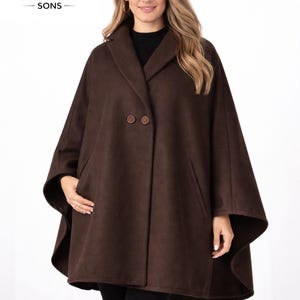 Op de afbeelding: Een bruine capejas met een klassiek ontwerp. De jas heeft een brede kraag, twee grote knopen en zakken. Het merklogo "SARFARAZ SONS" is zichtbaar in de linkerbovenhoek. Het model draagt een zwarte top en een zwarte broek.