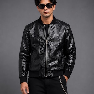 May include: A black bomber jacket with a textured, reptile-like pattern. The jacket features a front zipper, ribbed cuffs, and a ribbed hem. The person in the image is wearing black sunglasses and black pants.