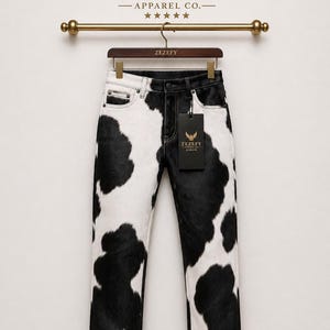 Peut inclure: Un jean à imprimé vache noir et blanc, suspendu à une tringle en bois. La marque "ZEZEFY" est visible. Le jean présente une coupe classique à cinq poches et un motif unique.