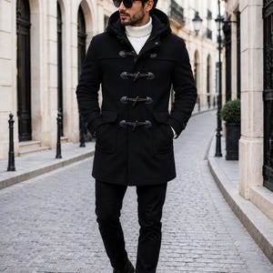 May include: A black hooded duffel coat with toggle closures, worn with black pants and boots. The coat has a classic design with a hood and large pockets. The outfit is completed with a white turtleneck and sunglasses.