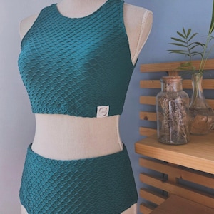 Puede incluir: Traje de baño de dos piezas de color verde azulado con un patrón texturizado en forma de gofre. La parte superior es un top corto con cuello alto y la parte inferior es de cintura alta. Una pequeña etiqueta blanca está cosida en la parte superior.