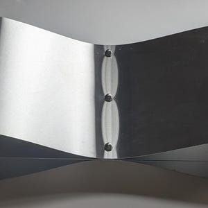 May include: A curved, brushed metal object with a bow-tie shape. The metal has a reflective surface and is held together by three small rivets. The object appears to be made of a single piece of metal.