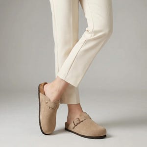 May include: Tan suede clogs with a cork footbed and adjustable buckle straps. The open-back design features a rounded toe and a dark brown sole. The clogs are shown with cream-colored pants.