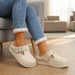 May include: Cream-colored slip-on clogs with a faux fur lining and a buckle strap. The clogs have a thick, white sole and are worn with blue jeans. The interior of the clogs is lined with a soft, fluffy material.