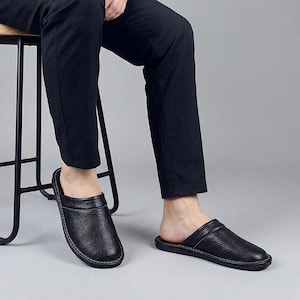 May include: Black leather slippers with a textured surface and white stitching. The slippers have a slip-on design and a rounded toe. The image shows a person wearing the slippers with black pants.