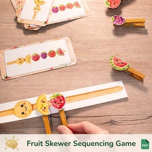 Op de afbeelding: Een houten tafel toont een fruitspies-sequentiegame. De game bevat kaarten en houten wasknijpers met fruitillustraties. De tekst "Fruit Skewer Sequencing Game" staat onderaan. De game bevat illustraties van een lachende koekje, citroen, framboos en watermeloen.