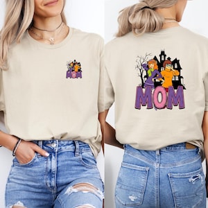 May include: Beige t-shirt with a graphic featuring cartoon characters and the word "MOM" in purple and pink. The front has a small graphic, while the back displays a larger design with a haunted house and characters.