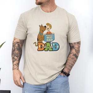 May include: Beige t-shirt featuring a cartoon graphic with the text "WORLD'S BEST DAD" on a blue sign held by a smiling cartoon character. A cartoon dog is kissing the character. The word "DAD" is in orange, green, and blue.