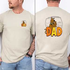 May include: Beige t-shirt featuring a cartoon image of a brown dog and the word "DAD" in orange and yellow. The front of the shirt has a smaller version of the graphic. The back of the shirt shows the dog in a van.