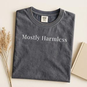 Könnte beinhalten: Ein dunkelgraues T-Shirt mit dem weißen Aufdruck "Mostly Harmless". Das T-Shirt liegt gefaltet auf einer cremefarbenen Oberfläche, neben einem beigen Buch und einem Zweig getrockneter Blumen. Das Shirt hat ein Comfort Colors-Etikett.