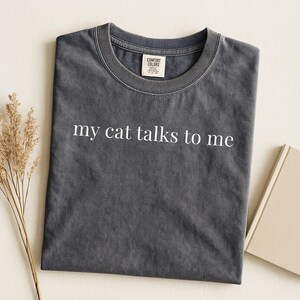 May include: Dark grey Comfort Colors t-shirt with the text "my cat talks to me" in white. The t-shirt is folded and placed beside a beige book and a sprig of dried flowers. The t-shirt has a crew neck.