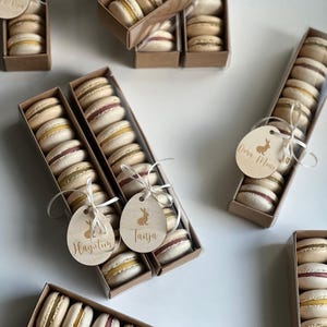 May include: Several boxes of colorful macarons, each containing multiple cookies. The macarons are in shades of yellow, pink, and white. Each box is adorned with a wooden tag featuring a bunny and a name. The boxes are arranged on a white surface.