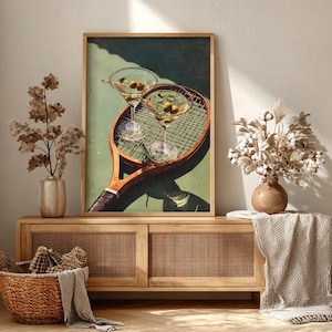 May include: A framed print depicting two martini glasses with olives resting on a vintage tennis racket. The artwork is displayed on a wooden cabinet with a woven front, with vases and a basket.