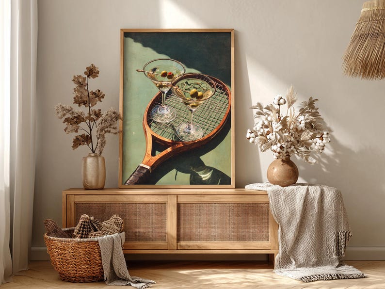 May include: A framed print depicting two martini glasses with olives resting on a vintage tennis racket. The artwork is displayed on a wooden cabinet with a woven front, with vases and a basket.